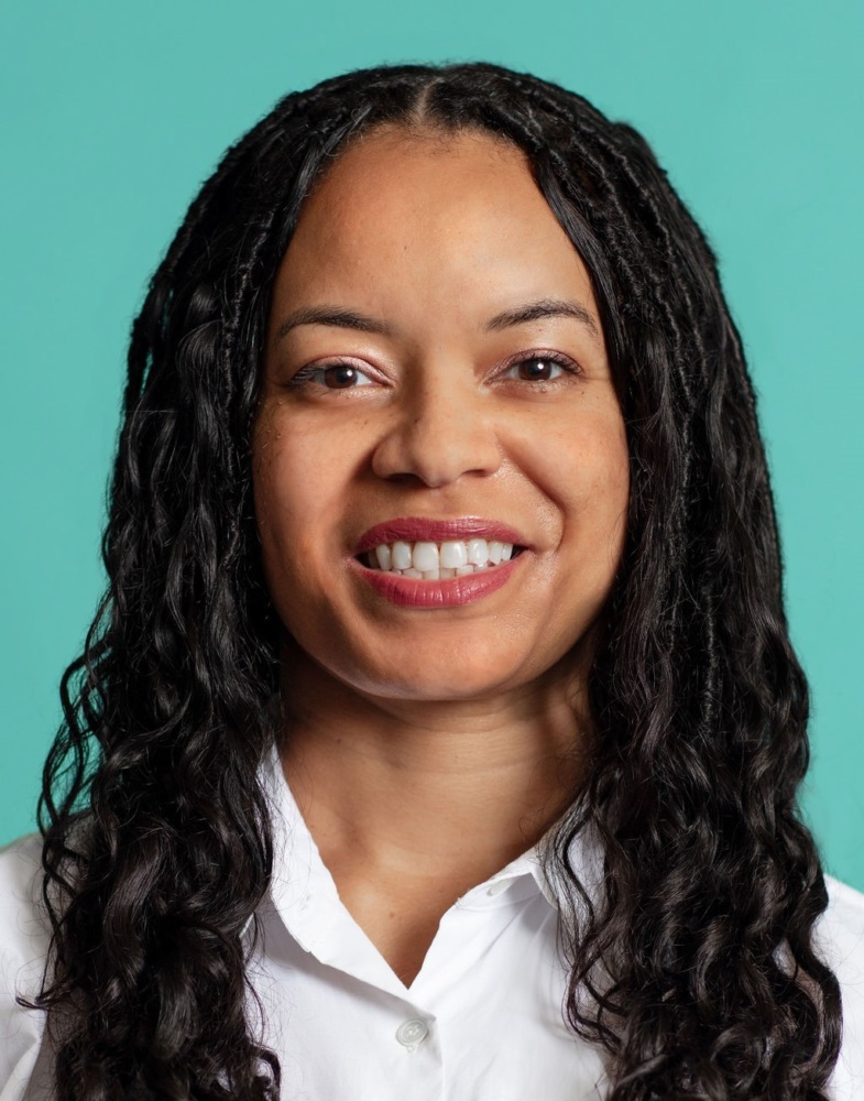 A woman with long curly hair is smiling in front of a plain turquoise background, wearing a white shirt.